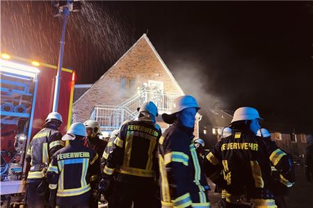 Zwei Feuerwehrleute mit Atemschutzgeräten bereiten das Betreten des Gebäudes vor, während ihre Kameraden an der Straße ihre Einsätze vorbereiten. 